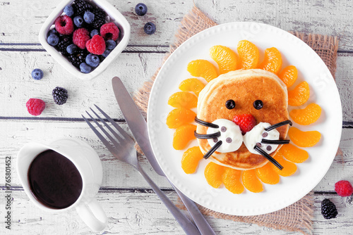 Cute child theme breakfast pancake in the shape of a lion face. Overhead view table scene on a white wood background.