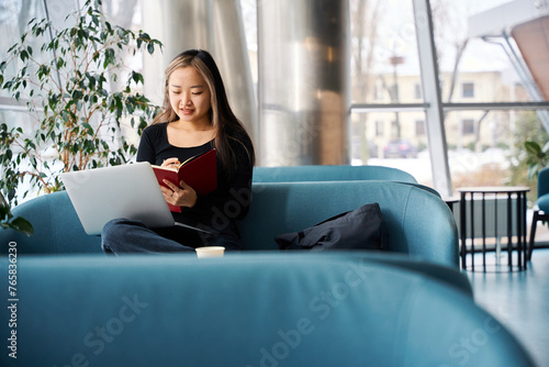 Young concentrated asian female IT employee with open laptop writing in notebook