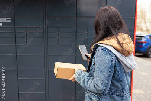 Young woman using smart phone while standing with a parcel delivered with post office machine with automatic lockers. New technologies in delivery service, self picking. photo