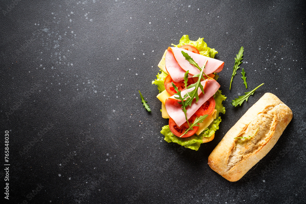 Ciabatta sandwich with lettuce, cheese, tomatoes and ham on black table.