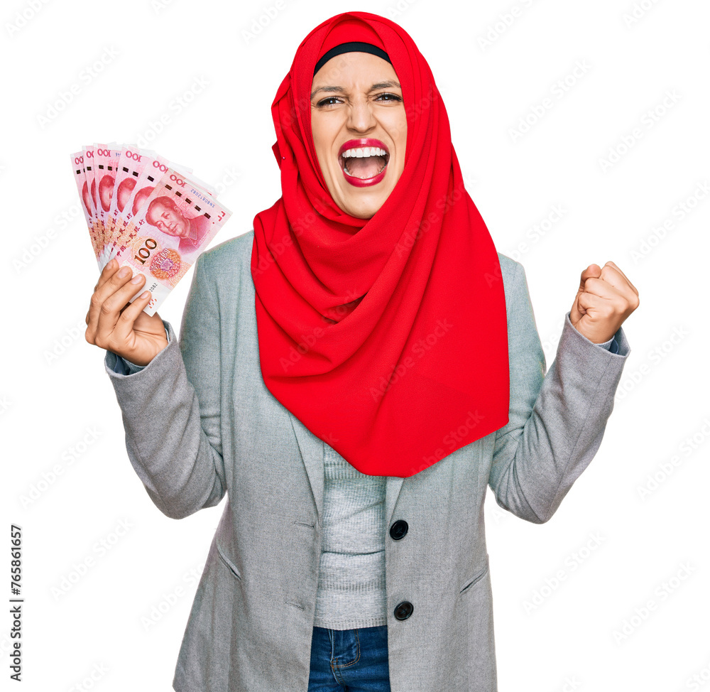 Beautiful hispanic woman wearing islamic hijab holding chinese yuan banknotes screaming proud, celebrating victory and success very excited with raised arms Beautiful hispanic woman wearing islamic hijab holding chinese yuan banknotes screaming proud, celebrating victory and success very excited with raised arms