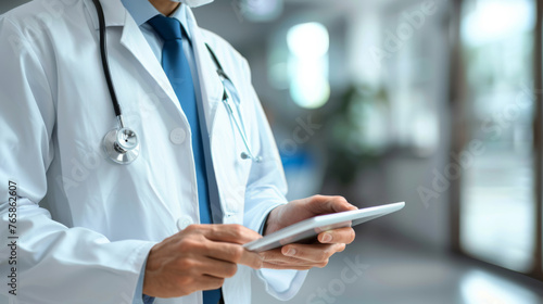A doctor in a white coat with a stethoscope is using a tablet in a hospital corridor.