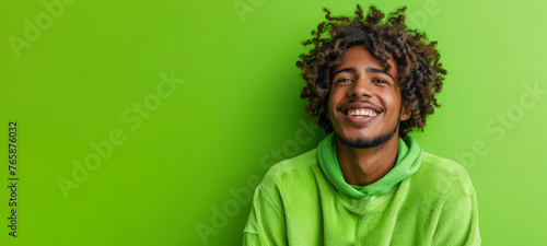 A man with curly hair is smiling and wearing a green hoodie. african american young man with curly hairstyle, smiling and laughing, wearing bright green clothes at bright solid green background