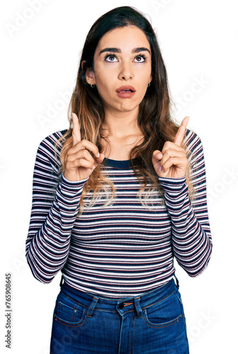 Beautiful hispanic woman wearing casual striped shirt amazed and surprised looking up and pointing with fingers and raised arms.