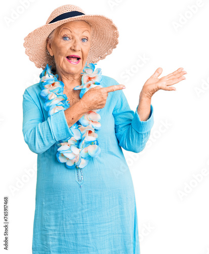 Senior beautiful woman with blue eyes and grey hair wearing summer hat and hawaiian lei amazed and smiling to the camera while presenting with hand and pointing with finger. © Krakenimages.com