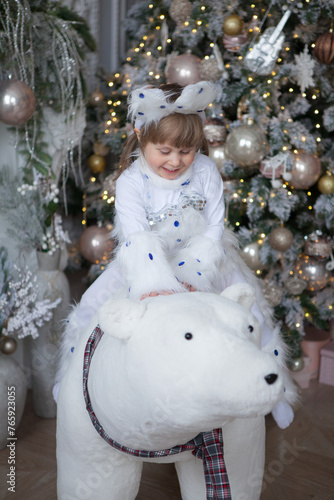 A little girl in a white cat costume is sitting on an artificial white bear by the Christmas tree.
