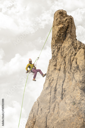 A man is climbing a rock wall with a green rope. The rope is attached to a harness on his back. The man is wearing a yellow jacket and purple pants. Heaven clouds.