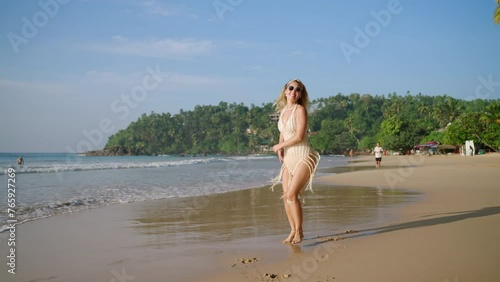 Happy young woman in swimwear enjoys freedom on tropical ocean beach, flirts, dances and finds pleasure in seaside relax by sea waves at exotic resort. Smiling young traveler blonde spins at shore. photo