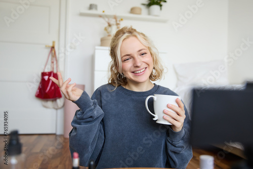 Portrait of cute smiling woman, blond girl drinks tea and records a casual, lifestyle video blog, vlogger sits in a room with camera and stabiliser, holds cup of coffee and talks photo