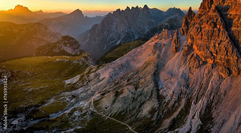 Drone panoramic shot of a hiking trail around Tre Cime di Lavaredo peaks in the Dolomites, with sharp summits in the background lit by the setting sun in vibrant orange-blue tones.