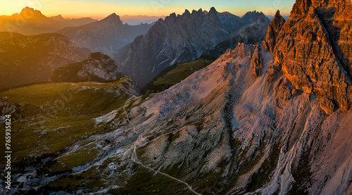 Drone panoramic shot of a hiking trail around Tre Cime di Lavaredo peaks in the Dolomites, with sharp summits in the background lit by the setting sun in vibrant orange-blue tones. © Martin Mecnarowski