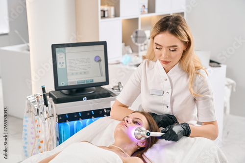 Female european beautician doing light therapy of face skin of young woman