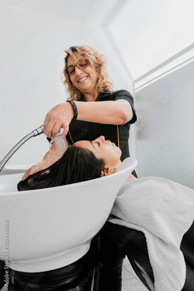 Positive hairstylist woman washing his client's brunette hair. White hairdresser salon interior. Hispanic client in a beauty salon. Positive hairstylist woman washing his client's brunette hair. White hairdresser salon interior. Hispanic client in a beauty salon.