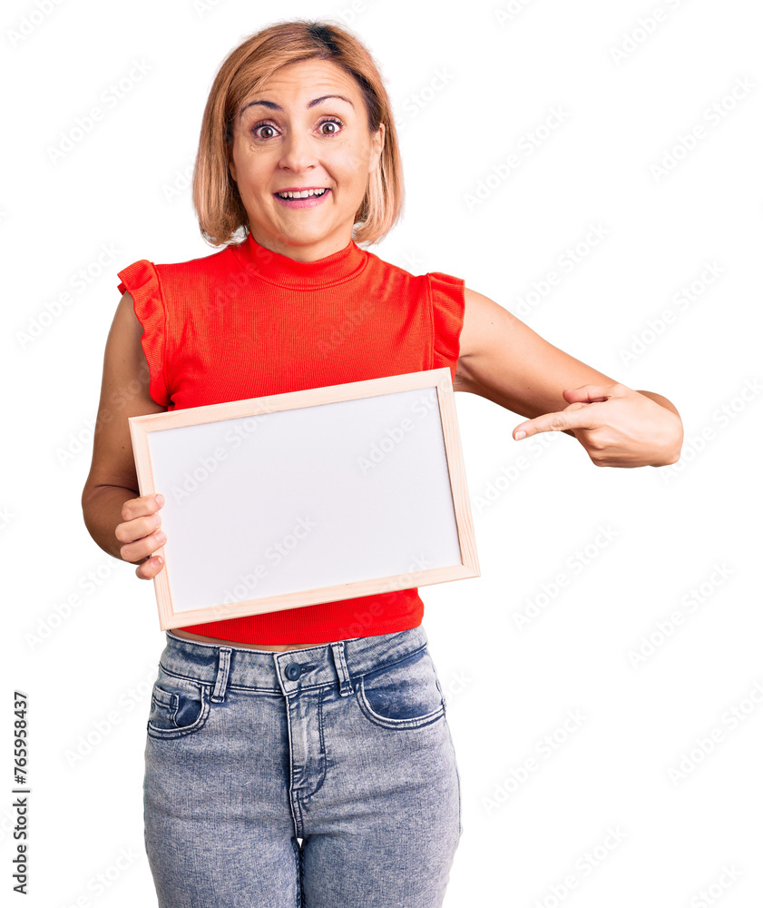 Young blonde woman holding empty white chalkboard smiling happy pointing with hand and finger Young blonde woman holding empty white chalkboard smiling happy pointing with hand and finger