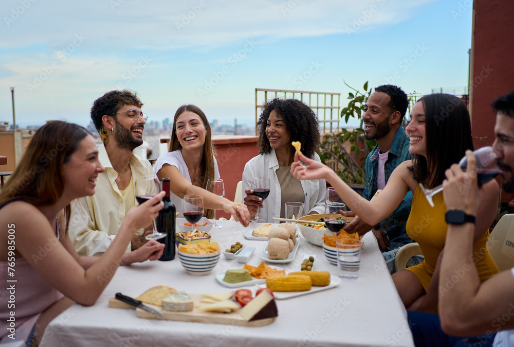 Laughing group of diverse young friends enjoying lunch together outdoors. Cheerful people gathered drinking red wine and eating snack on summer day having fun celebrating a birthday party on rooftop  - obrazy, fototapety, plakaty 