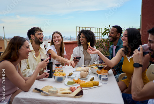 Laughing group of diverse young friends enjoying lunch together outdoors. Cheerful people gathered drinking red wine and eating snack on summer day having fun celebrating a birthday party on rooftop  photo