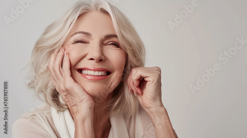 Joyful, radiant older woman with blonde hair gently touches her facial skin, gazing into the distance with a flawless smile, radiating laughter as she relishes in her mature beauty routine, contempla
