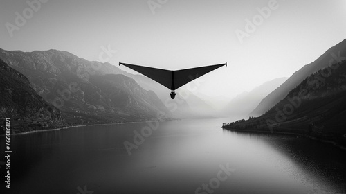 Black and white photo of a hang glider soaring above a mountain lake. 