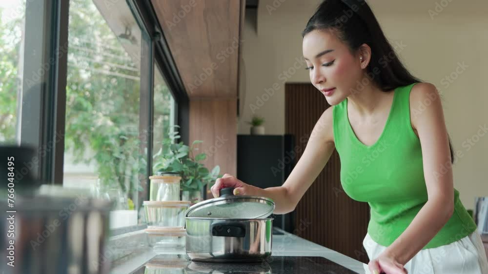 Happy Asian woman in green sleeveless open lid from steaming pot checking and smelling food cooking on a glass-top stove in kitchen Happy Asian woman in green sleeveless open lid from steaming pot checking and smelling food cooking on a glass-top stove in kitchen