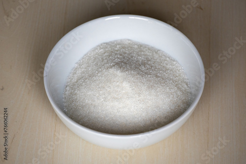 A white bowl is filled with sugar on a wooden textured background.