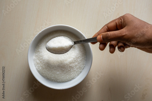 A white bowl is filled with sugar on wooden background. Woman's hand holding a sugar-filled spoon. Food ingredients for cooking sweets or desserts. photo