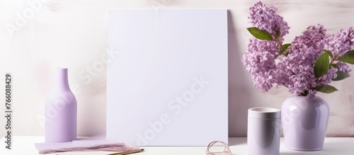 A white rectangle canvas is placed on a table alongside a magenta flower arrangement in a glass bottle filled with violet flowers and petals