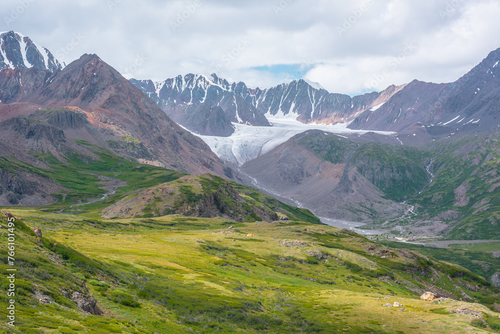 Colorful scenery of wide green alpine valley with view to large snow-capped range, sharp rocky pointy peak, snowy mountain range and big glacier tongue far away under cloudy sky in changeable weather.