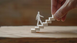 A hand is constructing a staircase with wooden blocks, with a paper cutout figure of a person stepping upwards, symbolizing growth and progress.