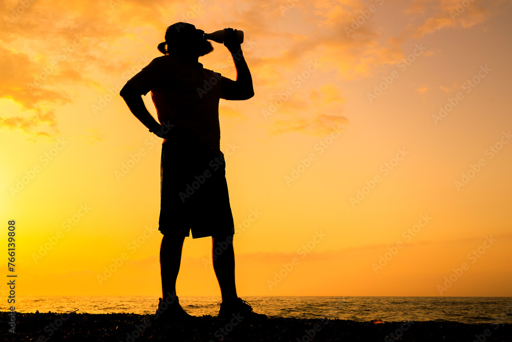 A man is drinking a beer while standing on a beach at sunset
