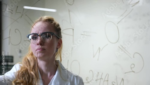A Caucasian woman in a medical gown thinks and finalizes formulas on a transparent wall.  photo