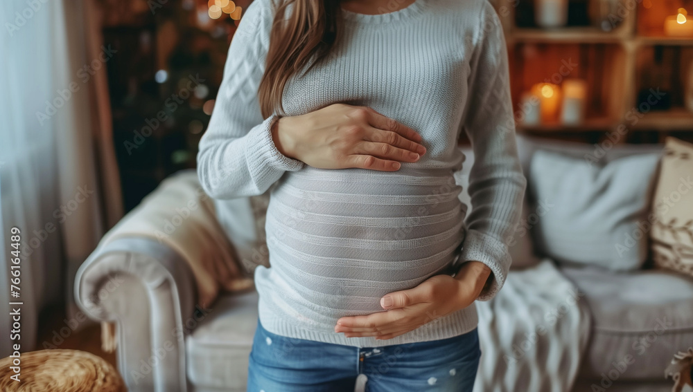 Close-up of a pregnant woman's belly, with hands holding her belly in a home environment, focusing on her hand and her round belly with a blurred background - obrazy, fototapety, plakaty 
