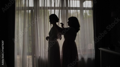 Silhouette of a mother combing her daughter's hair at the window. 
 Silhouette of mother braids her child against a window at sunrise.
