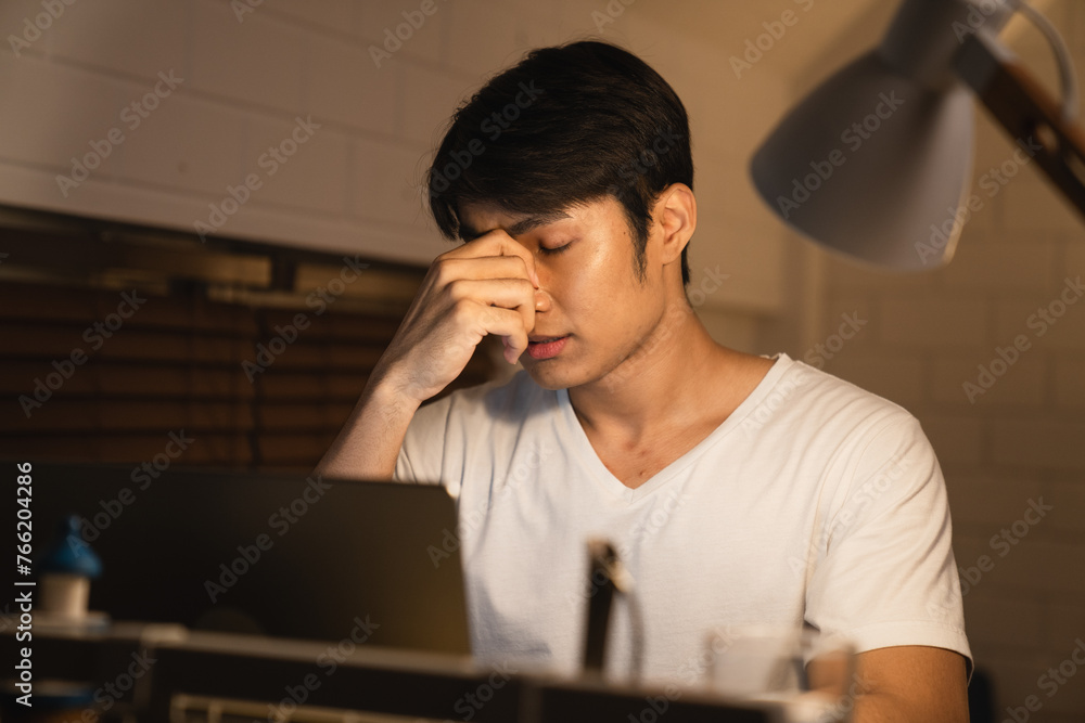 A stressed and sleepy Asian young man on a desk looking at a laptop computer in darkness late at night. Working. The feeling of hard work or hard work. A stressed and sleepy Asian young man on a desk looking at a laptop computer in darkness late at night. Working. The feeling of hard work or hard work.