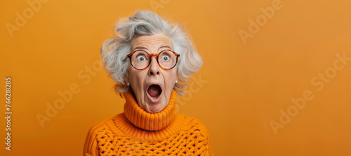 An older woman with glasses and a jacket is smiling and making a surprised face. Concept of joy. surprised happy funny old woman in glasses with mouth open on an orange background with a copy space