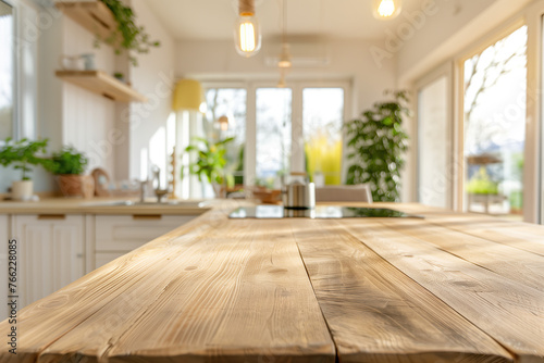 Blurred background of an empty wooden table in the foreground and modern kitchen with white cabinets, windows to sunny nature outside in the blurry backdrop. Concept for product display montage