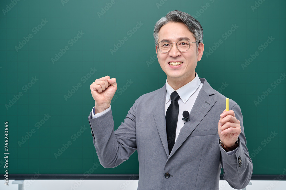 A middle-aged male teacher in a suit and glasses is giving an Internet lecture with a confident expression and pose with chalk in front of a green blackboard.
