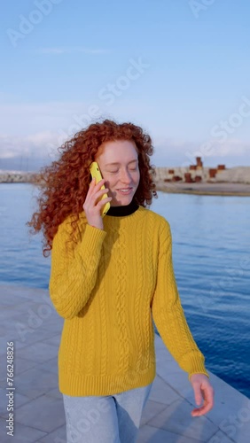 Redhead young woman talking on phone outdoors. Happy teenager walking at seaside.