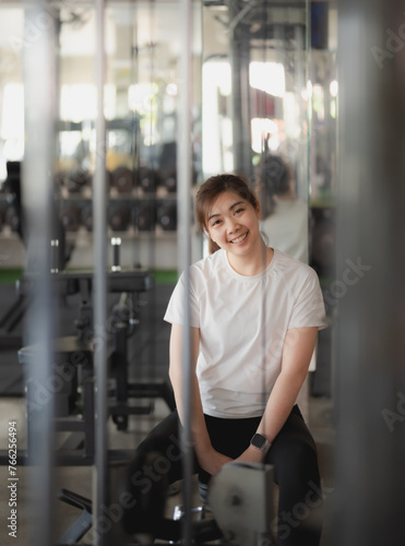 Asian sport woman is sitting on a weight machine with her arms crossed at fitness gym. She is smiling and looking at the camera. Fitness gym sport concept.