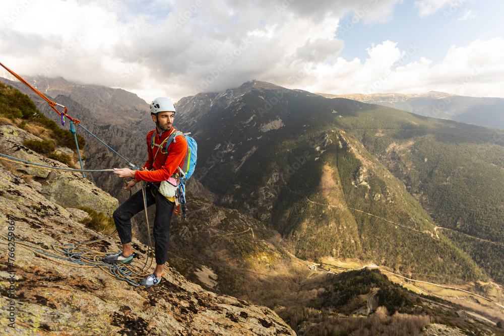 A man in a red jacket is standing on a rock with a rope attached to him. He is wearing a backpack and he is preparing for a climb. Concept of adventure and excitement