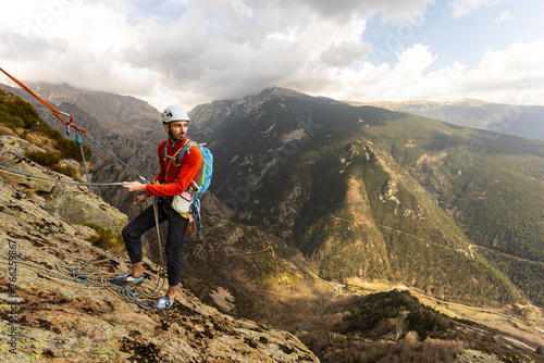 A man in a red jacket is standing on a rock with a rope attached to him. He is wearing a backpack and he is preparing for a climb. Concept of adventure and excitement