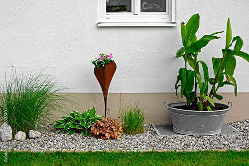 rośliny ozdobne w żwirowym ogrodzie obok domu, banan w doniczce, dekoracja z blachy corten, ogrodowa biżuteria z ordzewinego metalu, metal pot with a tropical plant, decoration in front of the house.