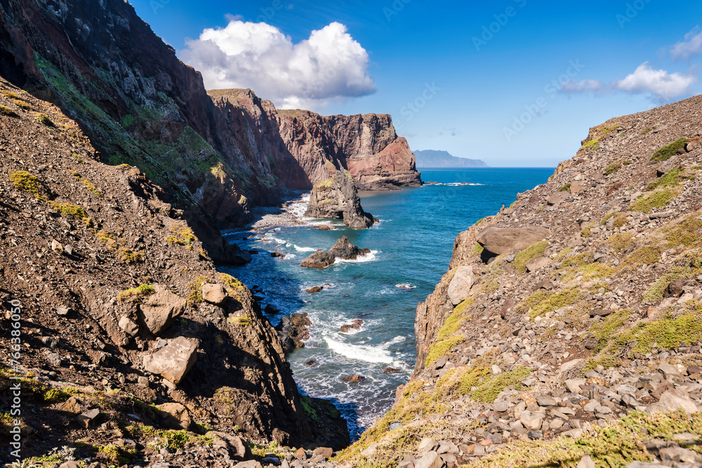 The photo depicts the rocky coast of Madeira. You can see the light blue sea, waves crashing against the rocks, and green vegetation on the slopes. It's a beautiful view of nature.