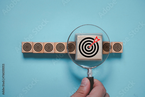 Hand holding magnifier glass to focus dartboard with arrow for business achievement objective goal and target concept. © Dilok