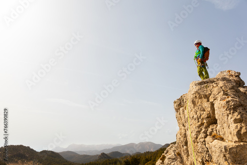A person is standing on a mountain top, looking out over the landscape. The sky is clear and the sun is shining brightly, creating a sense of peace and tranquility. The person is wearing a backpack