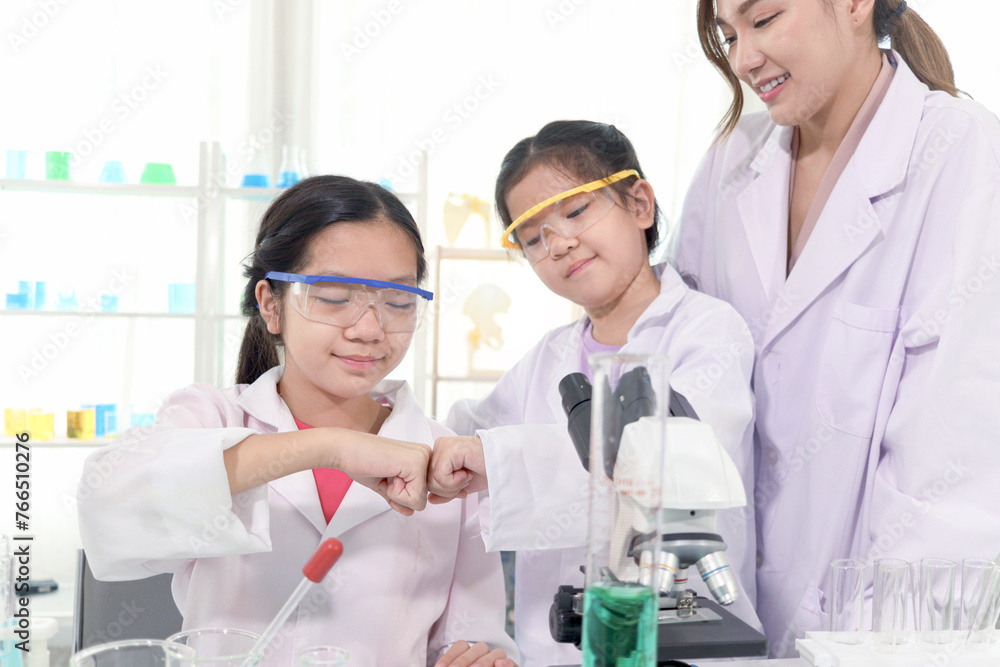 Young Asian student schoolgirls in lab coat bumping fists together to celebrate success of science experiment at laboratory. Student with female scientist teacher. Kid learning science education. Young Asian student schoolgirls in lab coat bumping fists together to celebrate success of science experiment at laboratory. Student with female scientist teacher. Kid learning science education.