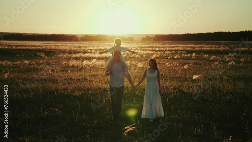 Father carries daughter on shoulders holding hand of wife walking in field. Family gets home together to celebrate mum birthday. Family discusses plans for future telling funny jokes at sunset photo
