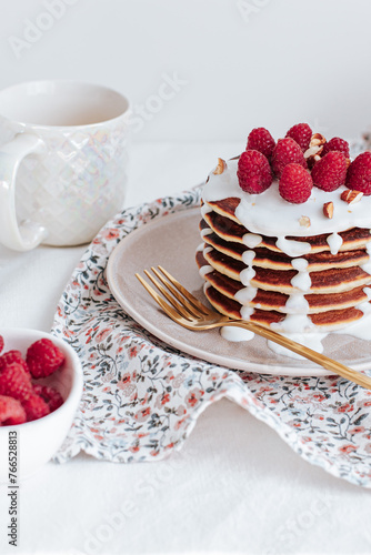 a stack of pancakes with vanilla sauce, decorated with raspberries and crushed almonds, behind a white cup with coffee and a bowl with raspberries. a colorful towel with gold cutlery. on a white backg
