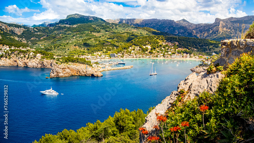 Sunlit panorama view of Port de Sóller embraced by the Serra de Tramuntana mountains, complemented by vibrant red flowers, a captivating Mediterranean getaway beckoning for exploration and relaxation.