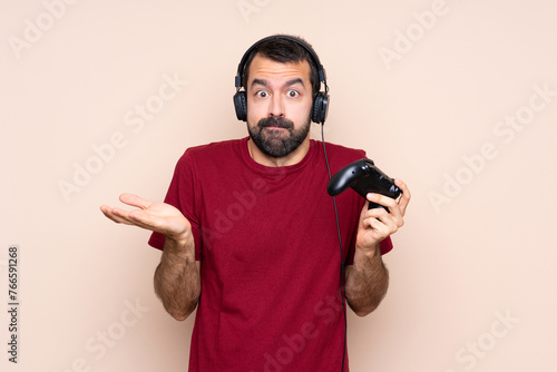 Man playing with a video game controller over isolated wall having doubts while raising hands