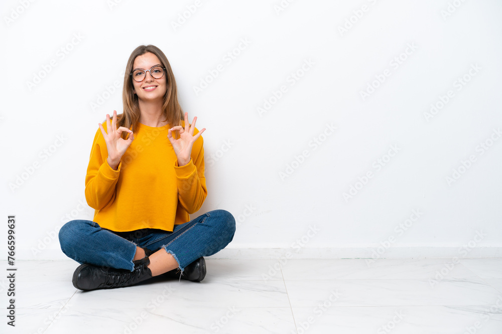 Young caucasian woman sitting on the floor isolated on white background showing an ok sign with fingers Young caucasian woman sitting on the floor isolated on white background showing an ok sign with fingers